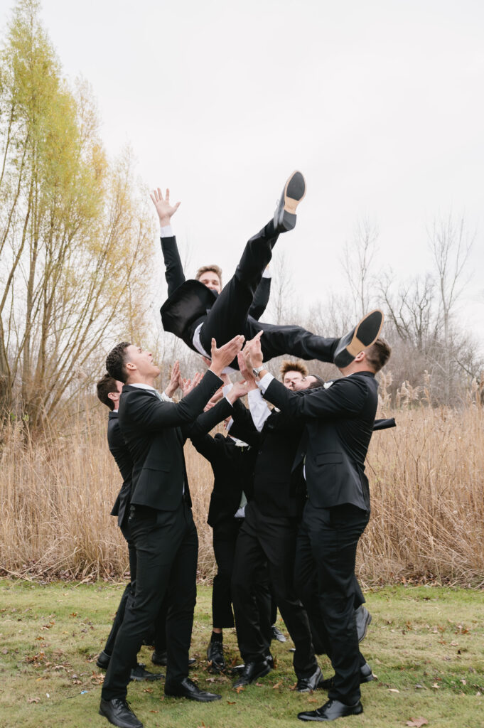 groomsmen tossing groom in open field near The Palazzo Grande, a wedding venue in Sterling Heights Michigan